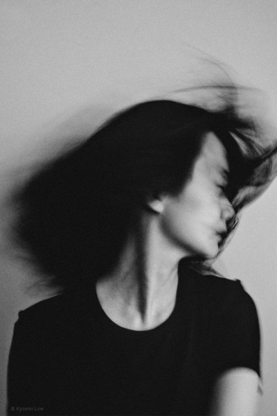 A black and white self-portrait of a woman in motion. Her face is turned to the side and softened by a deliberate motion blur from a slow shutter speed, creating a sense of fluid movement and quiet presence against a minimalist background.