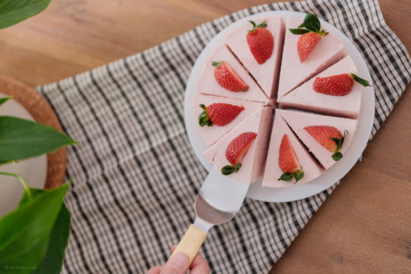 A close-up, top-down view of an egg-free strawberry bavarois on a white plate. A hand uses a silver cake server to lift a single slice, showing the smooth, mousse-like texture of the pink dessert and a garnish of fresh strawberries.