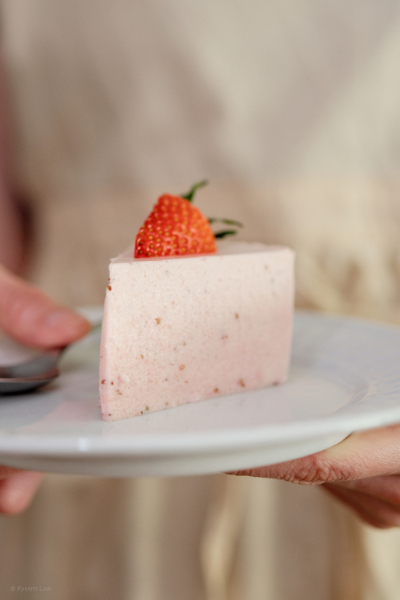202601_2_pastimes_kysemi_DSCF0835_web A side-profile view of a slice of pink strawberry bavarois sitting on a minimalist white plate. The image captures the clean, set edges of the dessert and the vibrant red of the fresh strawberry topping against a soft, neutral background.
