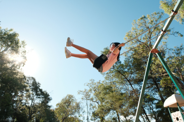 A low-angle silhouette of a young girl swinging high against a clear blue sky. The warm afternoon sun glows from the corner, highlighting her movement and the sense of freedom in a suburban park.
