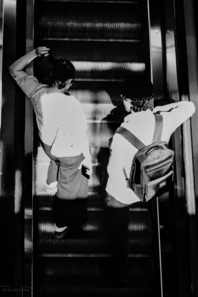 A high-contrast black and white street photograph from a high angle looking down an escalator. Two people are captured in a pool of bright afternoon light, creating strong geometric shadows and lines within a modern shopping center.