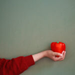 A minimalist, vertical shot of a person’s arm in a red sweater reaching into the frame, holding a bright red capsicum against a muted sage-green door. The image highlights the vibrant complementary colours.
