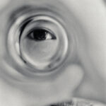 A close-up, artistic shot of a young girl's eye seen through a distorted drinking glass. The shallow depth of field creates a dreamlike effect, focusing on the details of the eye while the rest of the face is blurred.