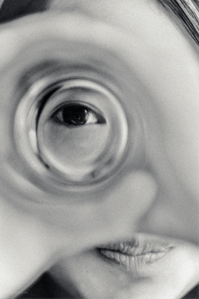 A close-up, artistic shot of a young girl's eye seen through a distorted drinking glass. The shallow depth of field creates a dreamlike effect, focusing on the details of the eye while the rest of the face is blurred.