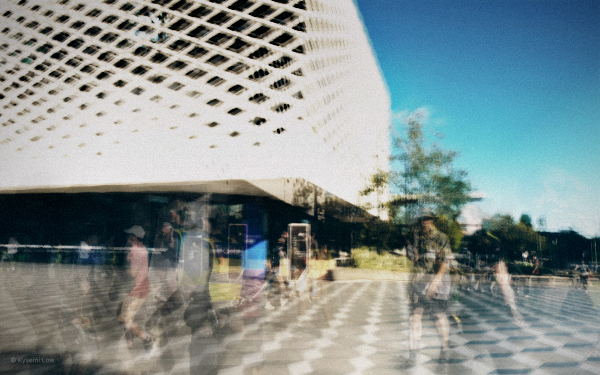 A multi-layered photograph of a bustling street. Multiple exposures of people and colours are layered together, creating a sense of movement that feels chaotic.