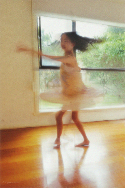 A motion-blur photograph of a young girl twirling in a floral dress in front of a bright window. The soft light and movement create a gentle, hazy atmosphere.