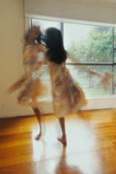 A painterly, long-exposure photograph of a young girl in a floral dress twirling in front of a window. Two images are layered together to create a stronger "double vision" effect, giving the movement a more artistic and painterly feel. 