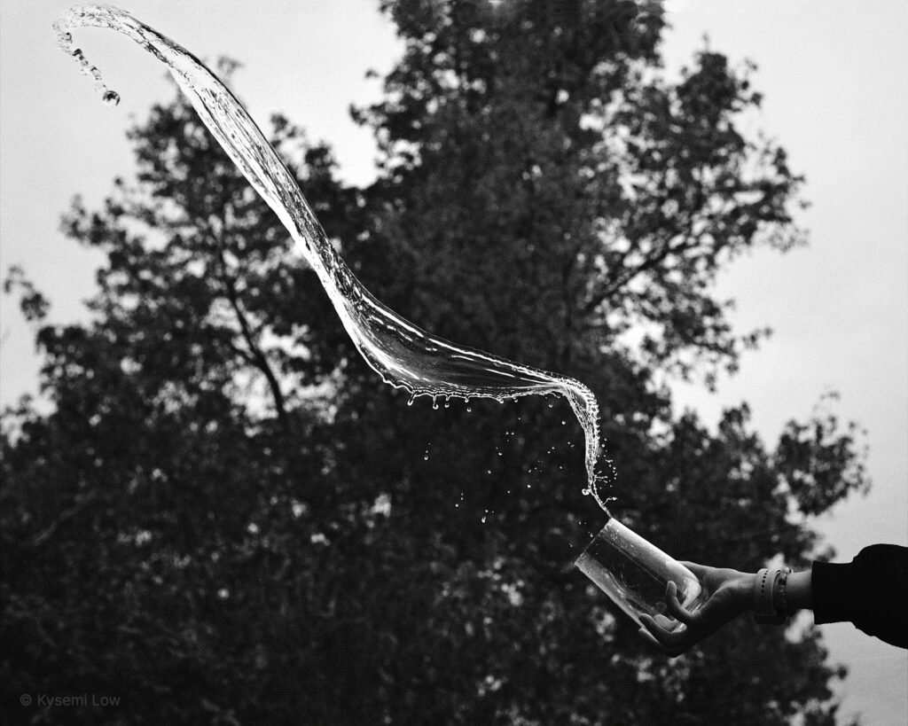 202605_21_water_kysemi_DSCF3007 A black and white photograph capturing dynamic movement: water flying outward in a bold, fluid wave from a tilted glass held by a steady hand, set against soft, dark trees.