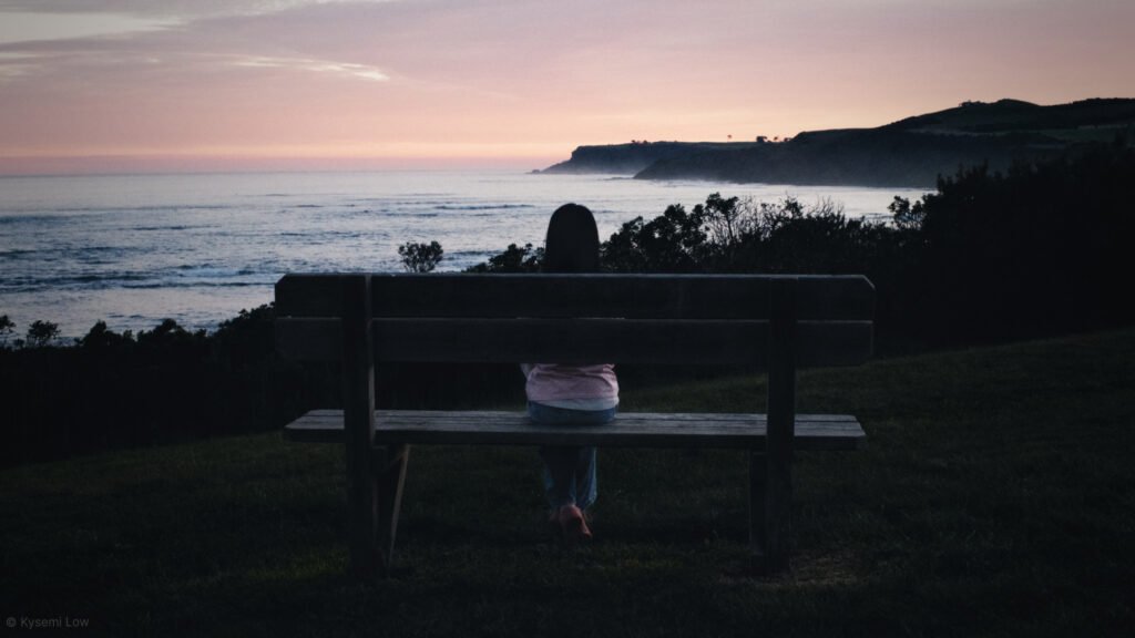 202606_28_inthecentre_kysemi_DSCF3782 A wide photograph at dusk showing a person sitting alone on a wooden bench on a grassy hill, looking out at the ocean horizon and distant dark hills.