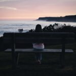 A wide photograph at dusk showing a person sitting alone on a wooden bench on a grassy hill, looking out at the ocean horizon and distant dark hills.