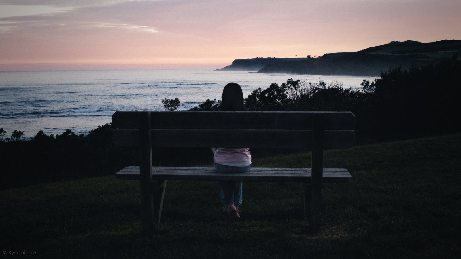 A wide photograph at dusk showing a person sitting alone on a wooden bench on a grassy hill, looking out at the ocean horizon and distant dark hills.